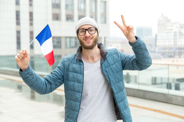 Happy man with a beard and glasses with the flag of France shows two fingers a V sign while standing against a city street.