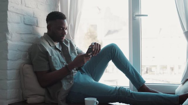 Handsome smiling african american young man relaxed sitting on windowsill in apartment with beautiful city views holding phone and surfing in the Internet