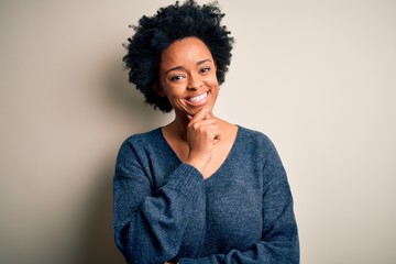 Young beautiful African American afro woman with curly hair wearing casual sweater looking confident at the camera smiling with crossed arms and hand raised on chin. Thinking positive.