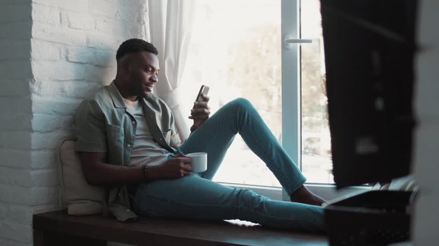 Handsome african american young man in the modern apartment sitting on windowsill looking through the window drinking cofee holding and using a smartphone