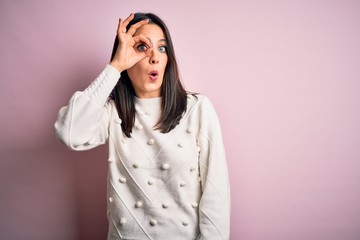 Young brunette woman with blue eyes wearing casual sweater over isolated pink background doing ok gesture shocked with surprised face, eye looking through fingers. Unbelieving expression.