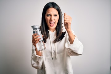 Young brunette sporty woman with blue eyes holding water bottle over white background annoyed and frustrated shouting with anger, crazy and yelling with raised hand, anger concept