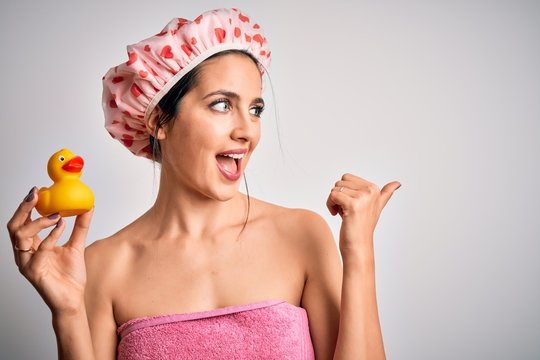 Young brunette woman with blue eyes wearing bath towel and shower cap holding duck toy pointing and showing with thumb up to the side with happy face smiling