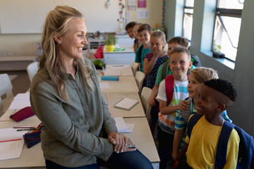 Female school teacher sitting on a desk in a classroom with a group of schoolchildren at elementary 