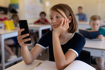 Schoolgirl sitting at a desk in an elementary school classroom using a smartphone