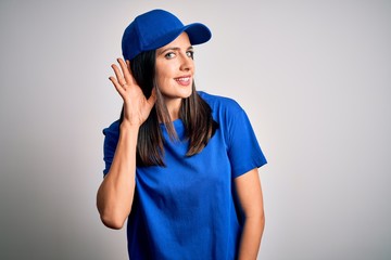 Young delivery woman with blue eyes wearing cap standing over blue background smiling with hand over ear listening an hearing to rumor or gossip. Deafness concept.