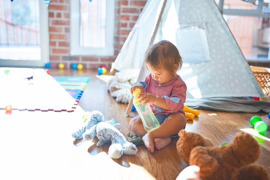 Adorable toddler holding feeding bottle around lots of toys at kindergarten