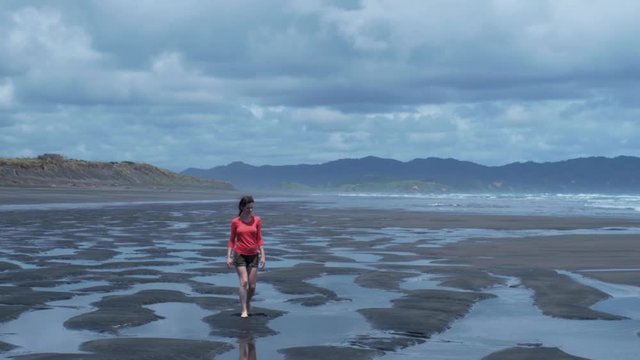 Young lonely woman walks leisurely through tidal pools on black sand beach on a cloudy day - Slow Motion