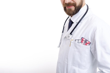 Cropped image of bearded doctor in smock with stethoscope and various pills and tablets in pocket isolated white background copyspace cropped