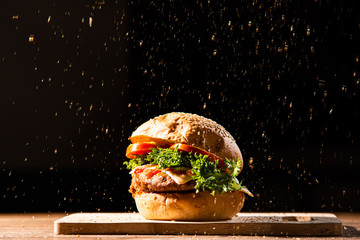home made hamburger made by white sesame bun, tomato slice, salad, cheese, grilled meat and onion on wooden tray wooden table with pouring oregano dark isolate background stock photo