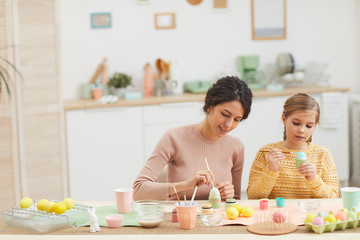 Obraz na płótnie Canvas Wide angle portrait of mother and daughter painting Easter eggs pastel color sitting at table in cozy kitchen interior, copy space