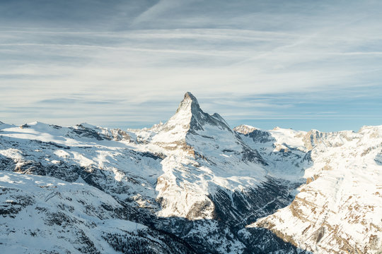 Scenic view on snowy Matterhorn (Cervin, Cervino) peak in sunny day with blue sky, pine trees, valley and clouds in background. Panoramic landscape in white and blue colors in Zermatt, Switzerland