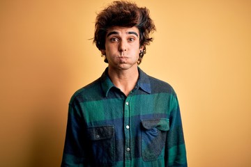 Young handsome man wearing casual shirt standing over isolated yellow background puffing cheeks with funny face. Mouth inflated with air, crazy expression.