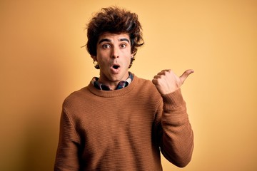 Young handsome man wearing casual shirt and sweater over isolated yellow background Surprised pointing with hand finger to the side, open mouth amazed expression.