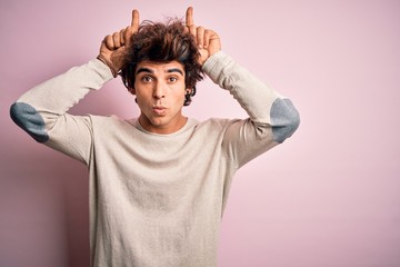 Young handsome man wearing casual t-shirt standing over isolated pink background doing funny gesture with finger over head as bull horns