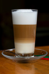 Hot latte coffee in tall translucent glass on wooden table on black background, vertical photography in low-key lightning.