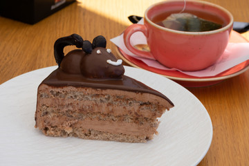 White plate with chocolate piece of cake with cream and edible chocolate mouse on top of it beside of cup of green tea.