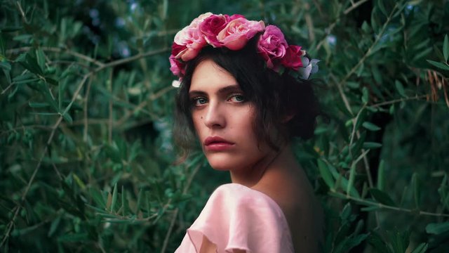 Cinemagraph / seamless video loop of a young caucasian female model / woman with a flower wreath and the wind moving her pink dress and hair and the green olive trees in the back.