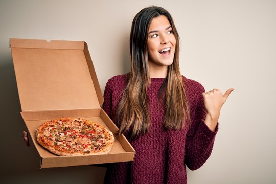 Young beautiful girl holding delivery box with Italian pizza standing over white background pointing and showing with thumb up to the side with happy face smiling