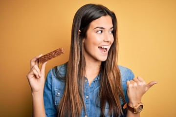 Young beautiful girl holding healthy protein bar standing over isolated yellow background pointing...