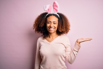 Young african american woman with afro hair wearing bunny ears over pink background smiling cheerful presenting and pointing with palm of hand looking at the camera.