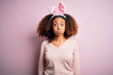 Young african american woman with afro hair wearing bunny ears over pink background puffing cheeks with funny face. Mouth inflated with air, crazy expression.