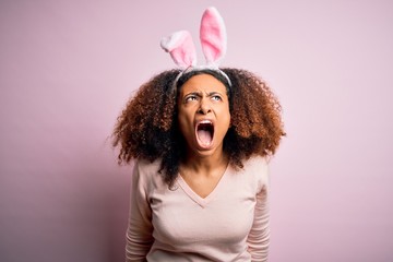 Young african american woman with afro hair wearing bunny ears over pink background angry and mad...