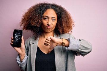 Young african american woman with afro hair holding cracked and broken smartphone screen with angry face, negative sign showing dislike with thumbs down, rejection concept