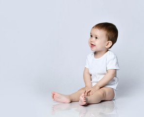 Happy smiling baby boy toddler in white bodysuit is sitting sideways on the floor looking up at copy space on a gray