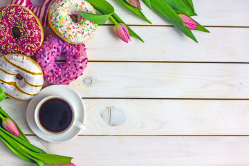Donuts background with coffee and flowers. Beautiful sweet donuts in multi-colored glaze, a cup of coffee, pink tulips on a wooden background, copy space, top view. Sweet background  holiday breakfast