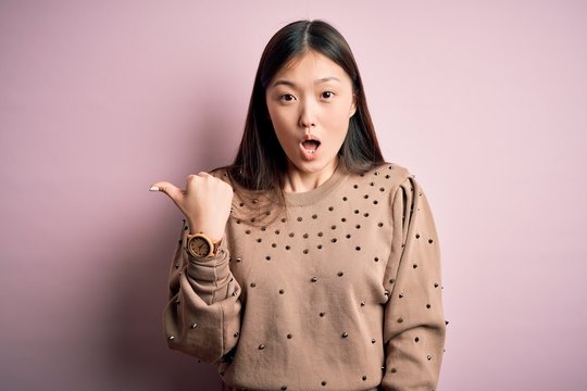 Young beautiful asian woman wearing fashion and elegant sweater over pink solated background Surprised pointing with hand finger to the side, open mouth amazed expression.