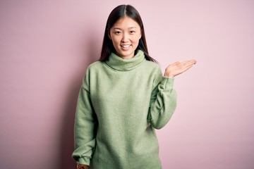 Fototapeta premium Young beautiful asian woman wearing green winter sweater over pink solated background smiling cheerful presenting and pointing with palm of hand looking at the camera.