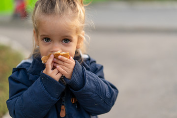A beautiful little Caucasian girl with blond hair and eating bread eagerly with her hands looks at...