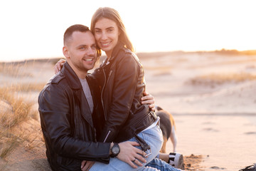 Beautiful young Caucasian couple dressed black leather jacket and blue jeans. A man sits on the sand woman sits on top and hugs him.