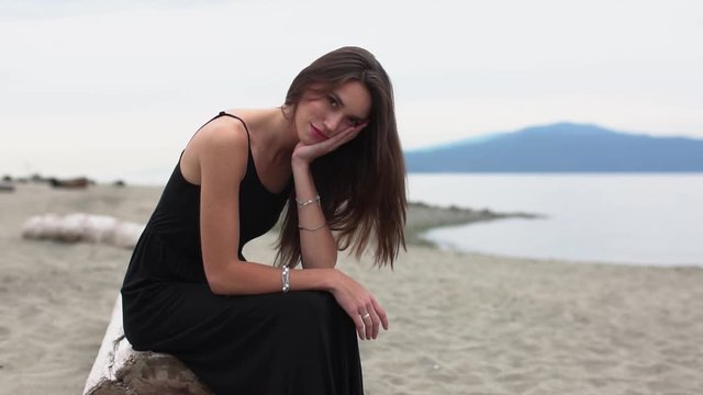 A closeup portrait video of a captivating woman in a black summer dress siting of a tree trunk washed away on the ocean shores. Slow-motion footage.