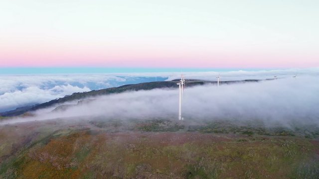 Drone footage of windmills on top of a mountain on the European island of Madeira, Portugal with white mist clouds rolling over the mountain top with a beautiful sunset gradient in the background