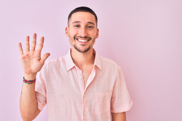 Young handsome man wearing pink shirt standing over isolated background showing and pointing up with fingers number five while smiling confident and happy.