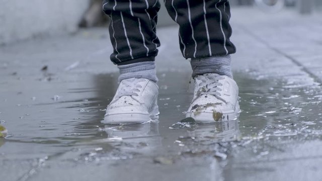 Child with white sneakers jumping into a puddle of rain water. cold wet winter 4k