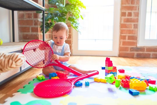 Beautiful toddler sitting on puzzle carpet holding tennis racket at kindergarten