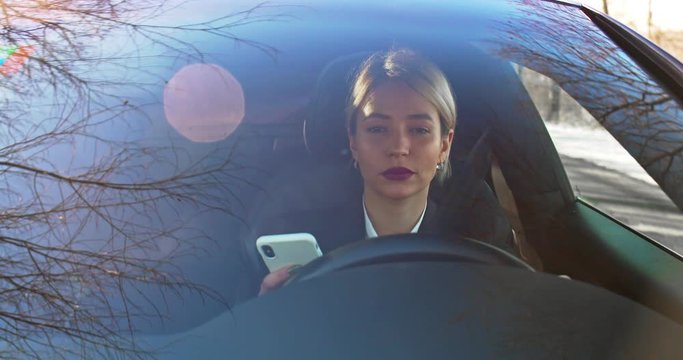 Caucasian young beautiful woman with fair hair tapping and texting on the smartphone while driving an expensive car.
