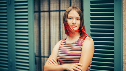 Outdoor portrait of pretty teenage girl with red dyed hair, wearing stripe vest, posing next to facade with green window