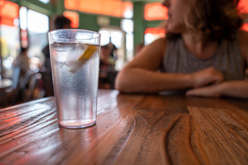 Glass of water with lemon and ice on restaurant table with woman