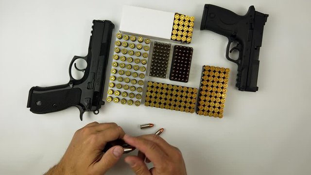 Overhead Dolly Shot Of a Man Removing  9mm Bullets from Handgun Magazine With Random boxes of ammunition and two pistols in the middle on the white background.