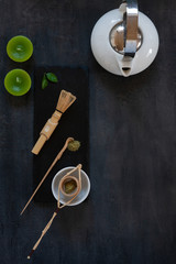 Matcha green tea ceremony set - white teapot, candles, matcha powder, wooden spoon, strainer and whisk on a dark gray background