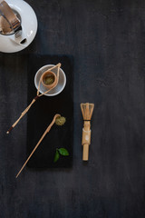 Matcha green tea ceremony set matcha powder, wooden spoon, strainer and whisk on a dark gray background.