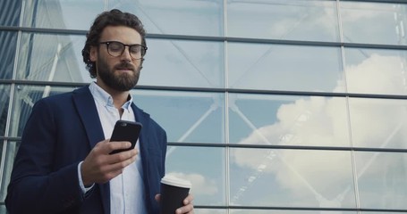 Handsome bearded Startup Businessman in Glasses using mobile Phone. Young man texting Messages, cheating on his Smartphone while walking in Office District. Business. Social Networking. People. Apps. - Powered by Adobe