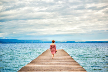 Back of faceless red haired woman in red dress walking by wooden pier on Garda lake in cloudy day. Italian vacation. Garda lake great tourist destination and attraction in North Italy.