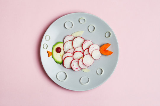 food art, creative children breakfast, cute funny fish made of colorful fresh tasty vegetables, carrot, zucchini, tomato, radish isolated on pastel pink background