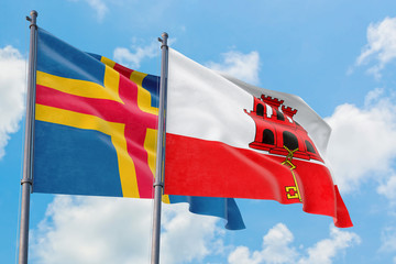 Gibraltar and Aland Islands flags waving in the wind against white cloudy blue sky together. Diplomacy concept, international relations.