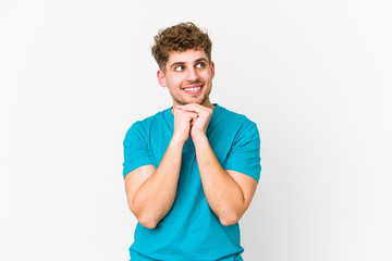 Young blond curly hair caucasian man isolated keeps hands under chin, is looking happily aside.